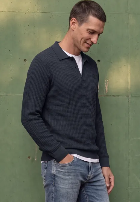 Man in donkerblauwe gebreide poloshirt met witte kraag en spijkerbroek, glimlachend voor een groene muur.