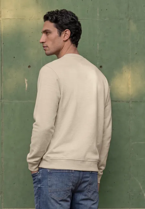 Ein Mann steht seitlich vor einer grünen Wand, trägt beige Sweatshirt und blaue Jeans.