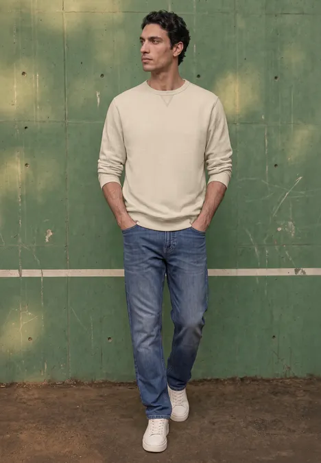 Ein Mann steht vor einer grünen Wand, trägt helles Langarmshirt, blaue Jeans und weiße Sneaker, Hände in den Taschen.