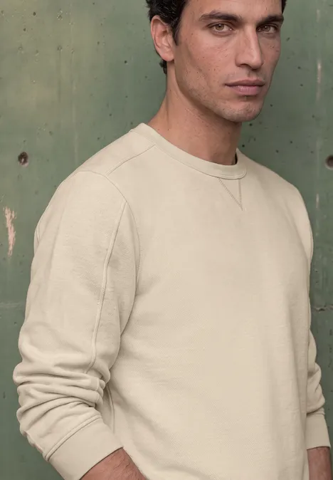 Junger Mann mit dunkelbraunen Haaren in beigem Sweatshirt steht vor einer grünen Wand.