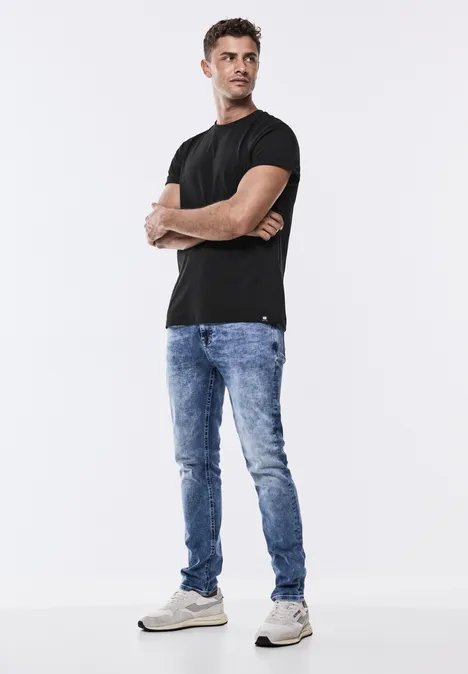 Mann mit schwarzem T-Shirt, blauer Jeans und weißen Sneakers steht mit verschränkten Armen.