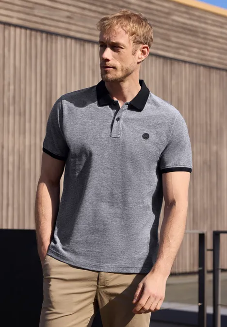 Un homme se tient devant un mur en bois, portant un polo gris à col noir et un pantalon beige.