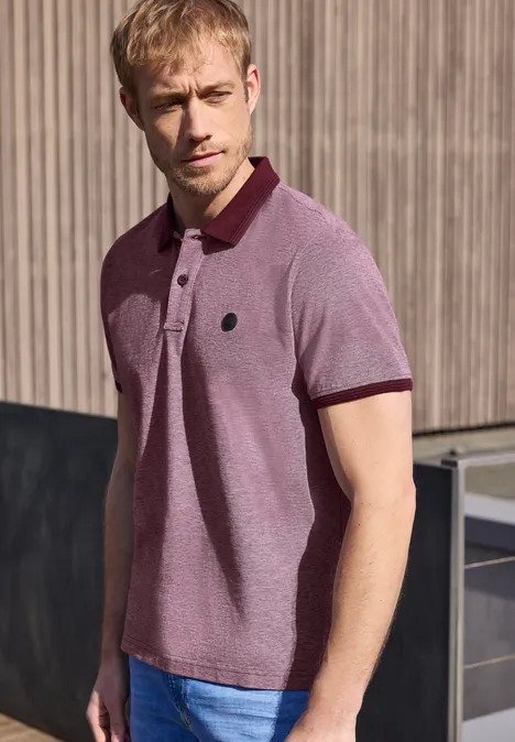 Un homme en polo rose se tient debout devant un mur en bois, regard tourné de côté.