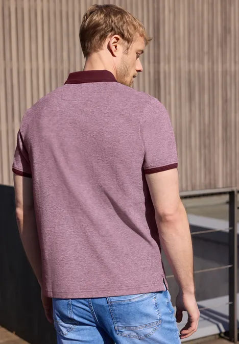 Un homme de dos porte un polo violet et un jean bleu devant une façade en bois.