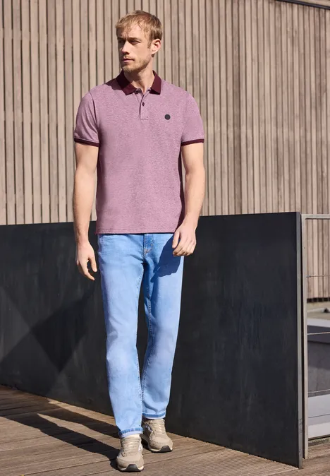 Un homme en jean et polo rose se tient devant une façade en bois.