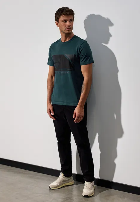 Mann steht an Wand, trägt grünes Shirt, schwarze Hose, helle Sneaker.