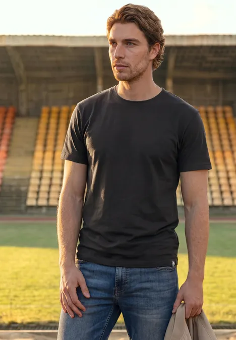 Basic Shirt in effen kleur Black Ein Mann in schwarzem T-Shirt und Jeans steht auf dem Rasen vor leeren Stadionrängen und blickt zur Seite.