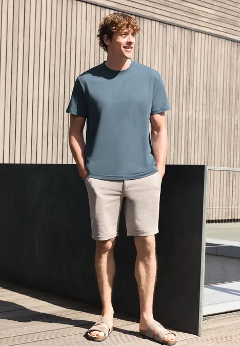 Ein junger Mann trägt ein blaues T-Shirt, beige Shorts und Sandalen, steht lässig vor einer Holzfassade.
