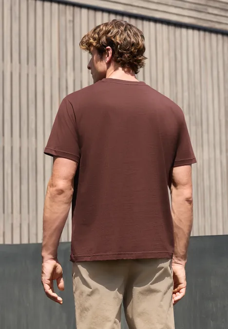 Ein Mann von hinten vor Holzfassade, braunes T-Shirt und beige Hose