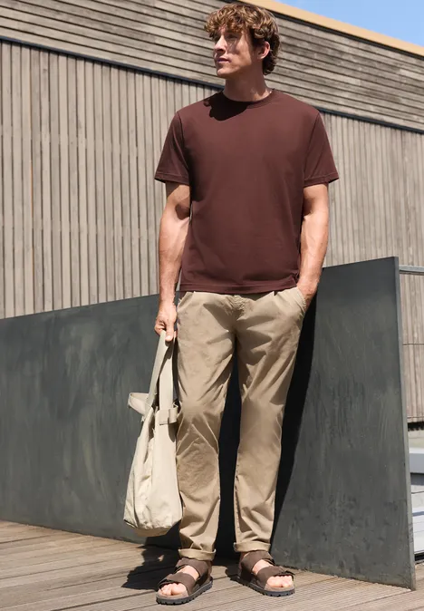 Ein Mann steht draußen vor einer Holzfassade, trägt braunes T-Shirt, helle Hose und Sandalen, hält eine Tasche.