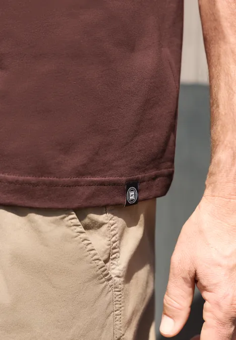 Nahaufnahme einer Person mit braunem T-Shirt und beigen Shorts, rechter Arm und Hand sichtbar.