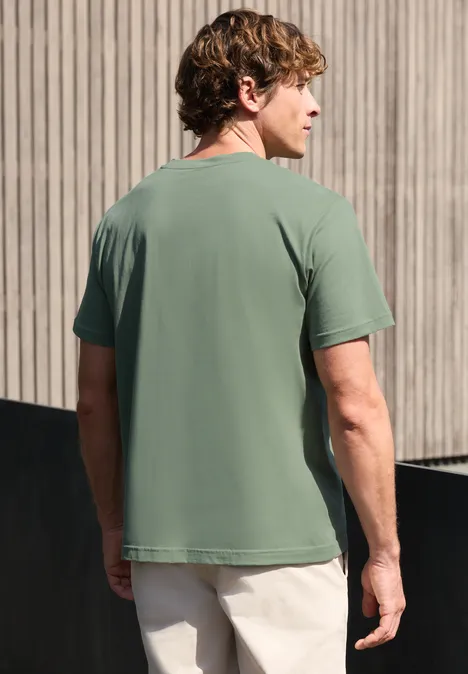 Ein Mann steht mit dem Rücken zur Kamera, trägt ein grünes T-Shirt und helle Hosen vor einer Holzfassade.
