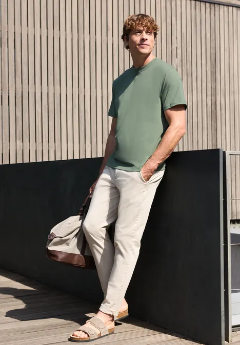 Ein Mann lehnt an einer Wand, trägt ein grünes T-Shirt, helle Hose, Sandalen und hält eine Reisetasche.