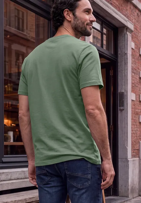 Ein Mann steht mit dem Rücken zur Kamera, trägt ein grünes T-Shirt und Jeans vor einem Gebäude.