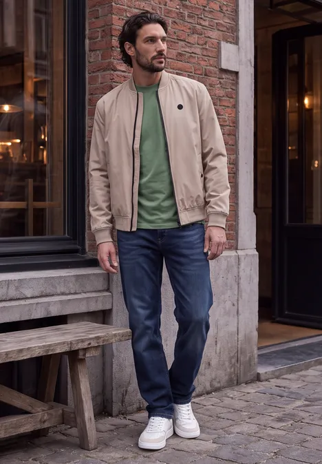 Ein Mann in beiger Jacke, grünem Shirt und Jeans steht vor einem Backsteingebäude auf dem Gehsteig.