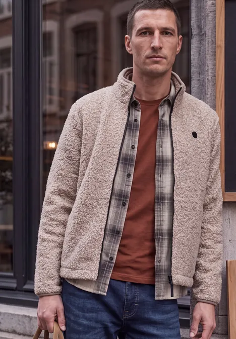 Mann in beiger Teddyjacke, kariertem Hemd und orangem Shirt vor Fensterfassade.