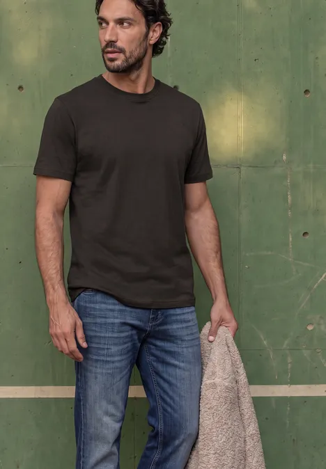 Mann vor grünem Hintergrund trägt dunkles T-Shirt, Jeans und hält eine beige Jacke.