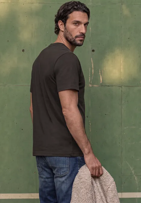 Ein bärtiger Mann in schwarzem T-Shirt und Jeans blickt über die Schulter vor grüner Wand und hält ein Tuch.
