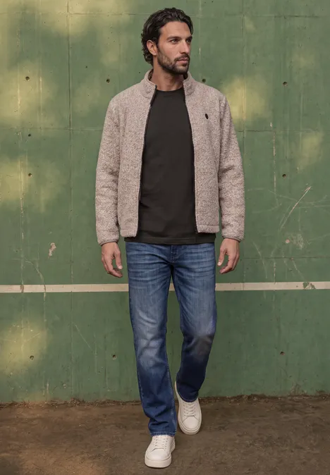 Ein Mann in Jeans, dunkelgrünem Pullover und beiger Jacke geht vor einer grünen Wand.