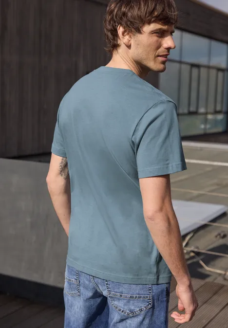 Ein Mann steht auf einer Terrasse und blickt über die Schulter, trägt ein türkis-blaues T-Shirt und Jeans.