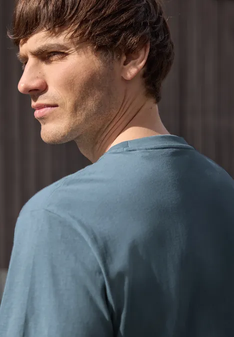 Ein junger Mann steht mit dem Rücken zur Kamera und schaut über seine Schulter, trägt ein blaues T-Shirt.
