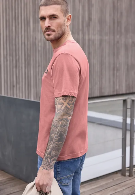 Ein tätowierter Mann in rosa T-Shirt und Jeans steht seitlich vor einer Betonwand mit Holzverkleidung.