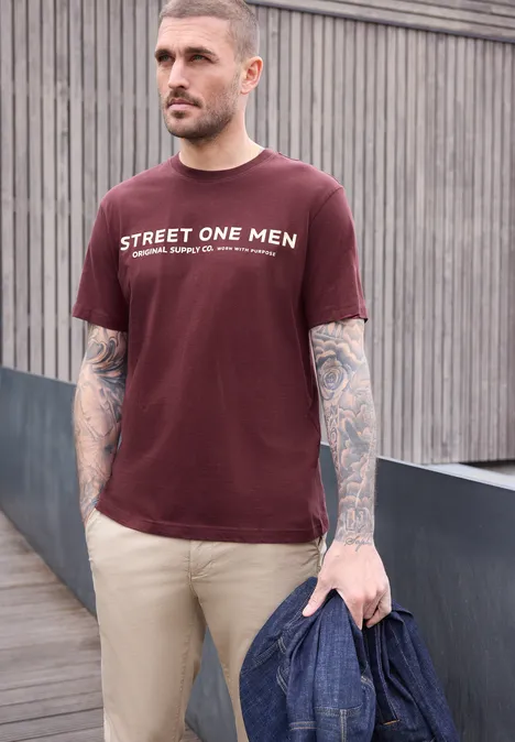 Ein tätowierter Mann trägt ein burgunderrotes T-Shirt und hält eine Jeansjacke.
