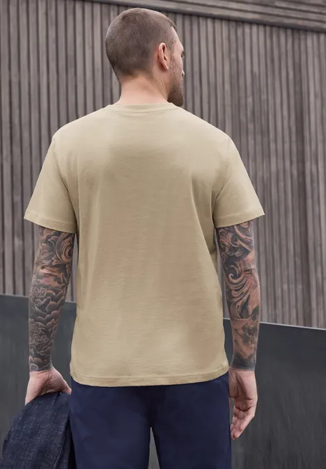 Ein tätowierter Mann steht mit dem Rücken zur Kamera vor einer Holzverkleidung, trägt ein beiges T-Shirt.