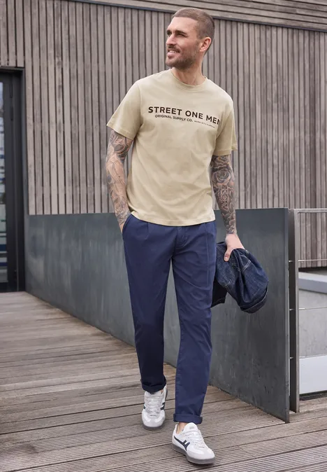 Ein tätowierter Mann in beige T-Shirt und blauen Hosen läuft mit Jacke in der Hand.