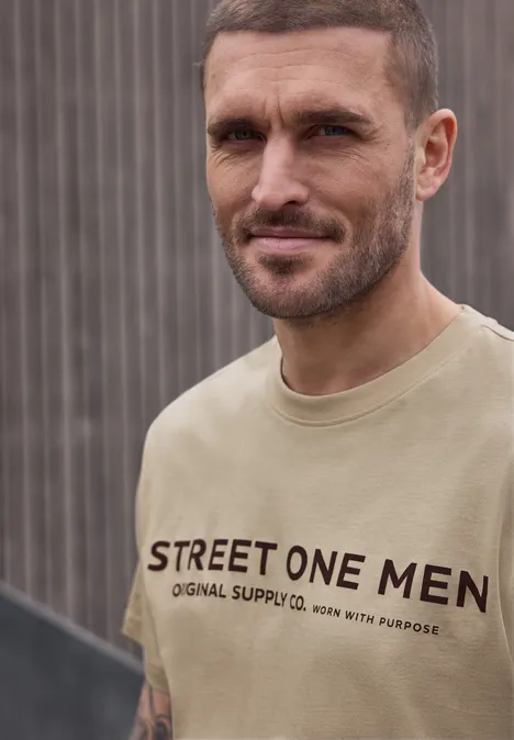 Ein verschwommenes Porträt eines Mannes in beige T-Shirt mit Aufdruck „STREET ONE MEN“ vor grauem Hintergrund.