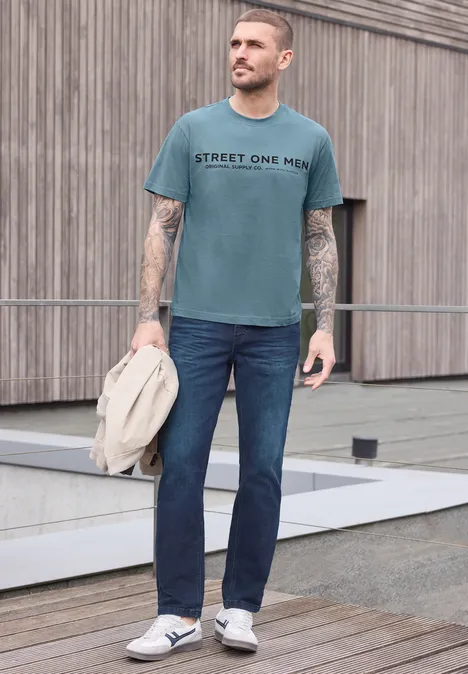 Mann in taubenblauem T-Shirt mit Aufschrift, Jeans und Sneakern steht vor Holzwand und hält Jacke.