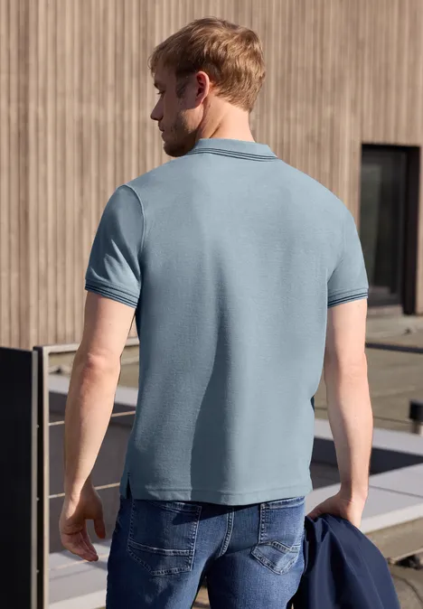 Un homme de dos porte un polo bleu clair et un jean, tient une veste devant une façade en bois.