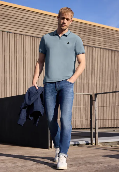 Un homme en polo bleu, jean et baskets se tient sur une terrasse en bois, tenant une veste à la main.