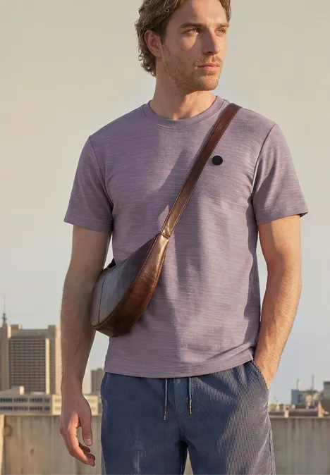 Mann in lila T-Shirt und brauner Umhängetasche steht auf sonnigem Dach mit Stadtblick.