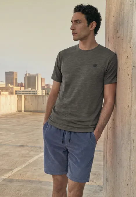 Ein Mann lehnt an einer Betonstütze auf einem Dach, trägt T-Shirt und Shorts.