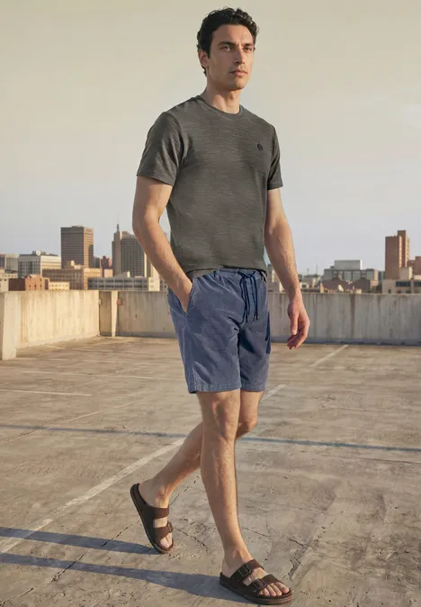 Cosy Slub T-Shirt mit Struktur oil grey Ein Mann steht auf einem Parkdach, trägt graues T-Shirt, blaue Shorts und Sandalen, Skyline im Hintergrund.