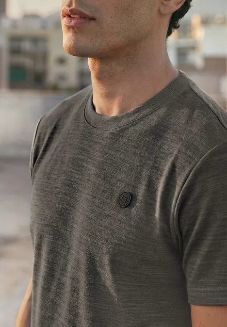 Cosy Slub T-Shirt mit Struktur oil grey Ein Mann trägt ein graues T-Shirt mit kleinem Brustlogo, unscharfer urbaner Hintergrund.