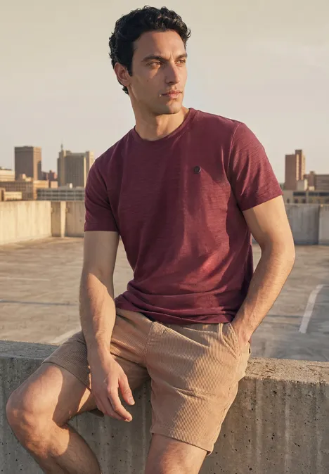 Ein Mann sitzt auf einer Dachkante, trägt rotes T-Shirt und beige Shorts, Blick in die Ferne.