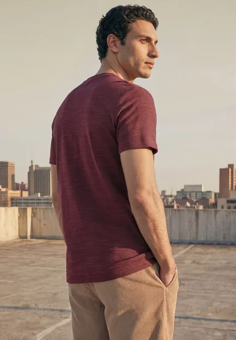 Een man staat op een dak, kijkt over de stad en draagt een roodbruin T-shirt met beige broek.