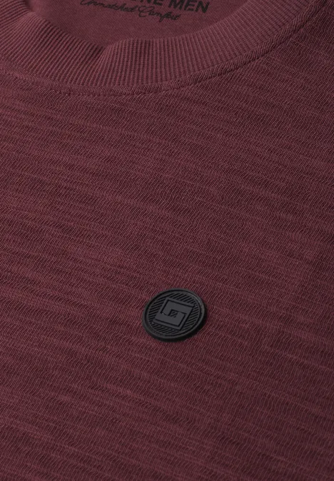 Bordeauxrood gebreid T-shirt met zwart rond logo-embleem op de borst.