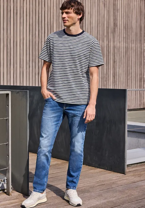 Ein junger Mann in gestreiftem T-Shirt und Jeans, steht auf einer Holzterrasse vor moderner Fassade.