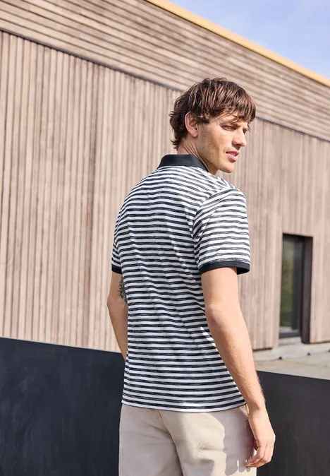 Ein junger Mann in gestreiftem Poloshirt und beigen Hosen steht mit abgewandtem Blick vor einer Holzwand.