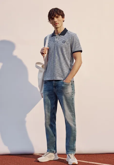 Junger Mann in gestreiftem Poloshirt, Jeans und Sneakers, stehend mit Tasche und Schatten im hellen Licht.
