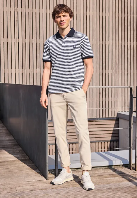 Ein Mann lehnt draußen an einer Holzstruktur und trägt ein gestreiftes Poloshirt, helle Hose und weiße Sneaker.