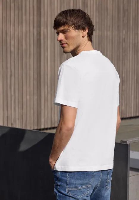 Ein junger Mann steht seitlich vor Holzwand, trägt weißes T-Shirt und blaue Jeans.