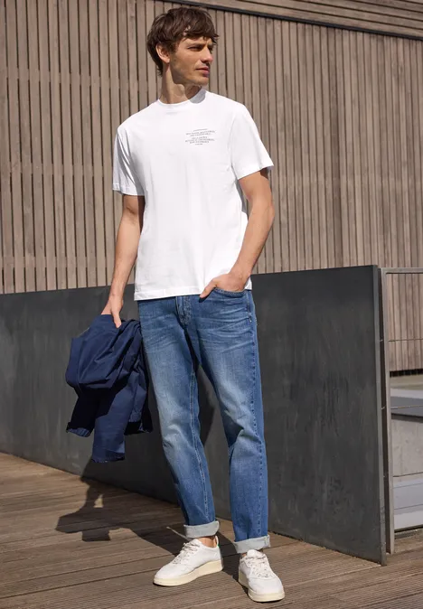Mann in weißem T-Shirt, Jeans und Sneakern hält dunkelblaue Jacke, steht lässig vor Holzfassade.