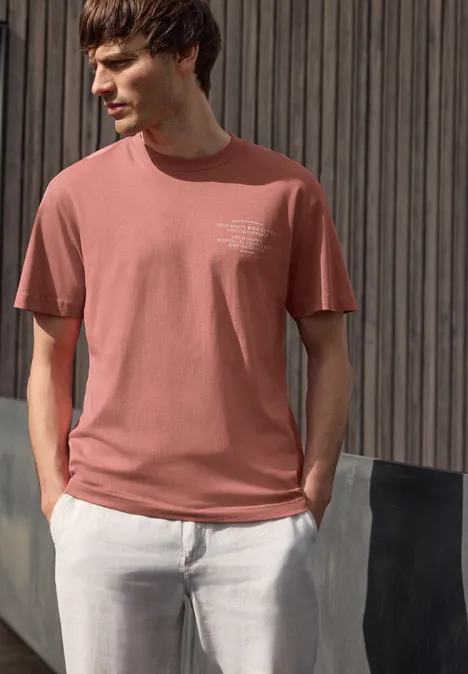 Mann in rosa T-Shirt und weißer Hose vor Holzwand.