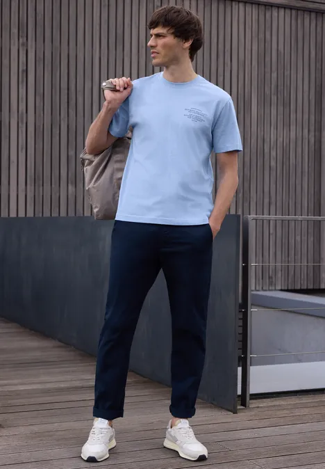 Ein Mann trägt lässiges Outfit mit hellblauem T-Shirt, dunkelblauen Hosen und weißen Sneakern draußen.