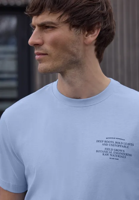 Ein seitlich abgebildeter Mann trägt ein hellblaues T-Shirt mit kleinem schwarzen Logo, unscharfes Gesicht.