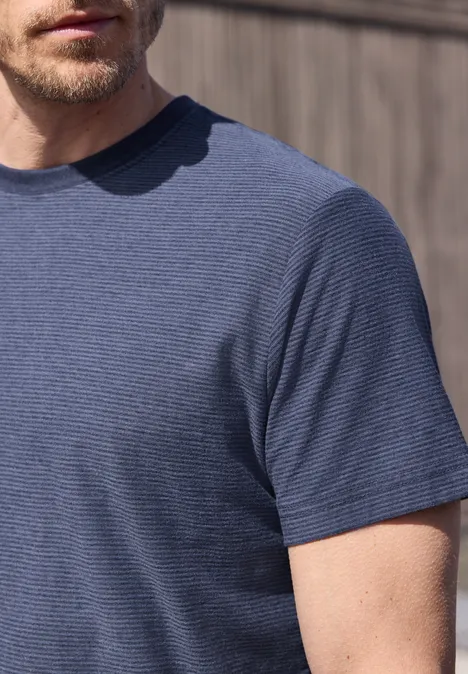 Ein Mann mit kurzem Bart trägt ein leicht gestreiftes blaues T-Shirt vor einer Holzwand.